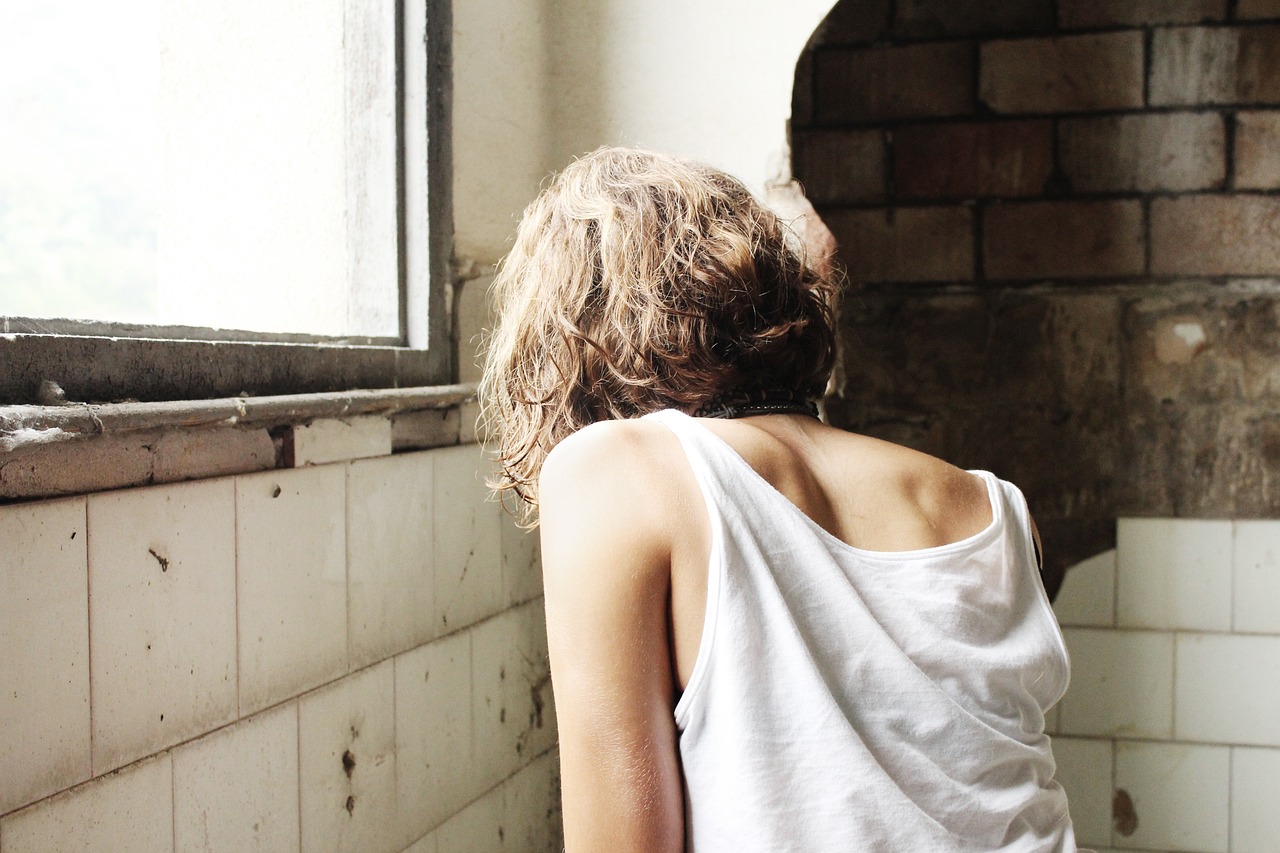  Image of a woman's back, wearing a white tank top, loose tile on wall, window frame is broken.